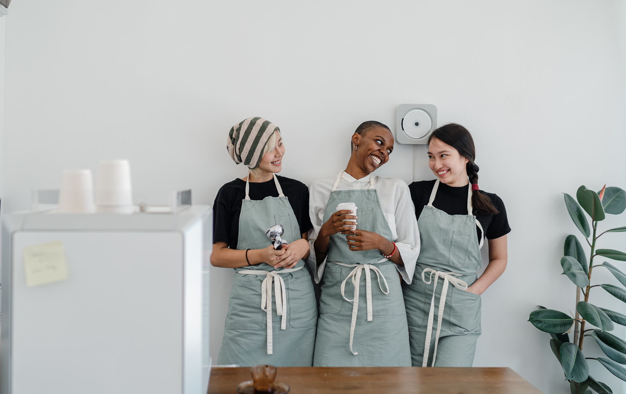 Image of 3 friends who work at the same cafe, wearing their aprons and smiling to eachother. The cafe has plants and a minimalist aesthetic.