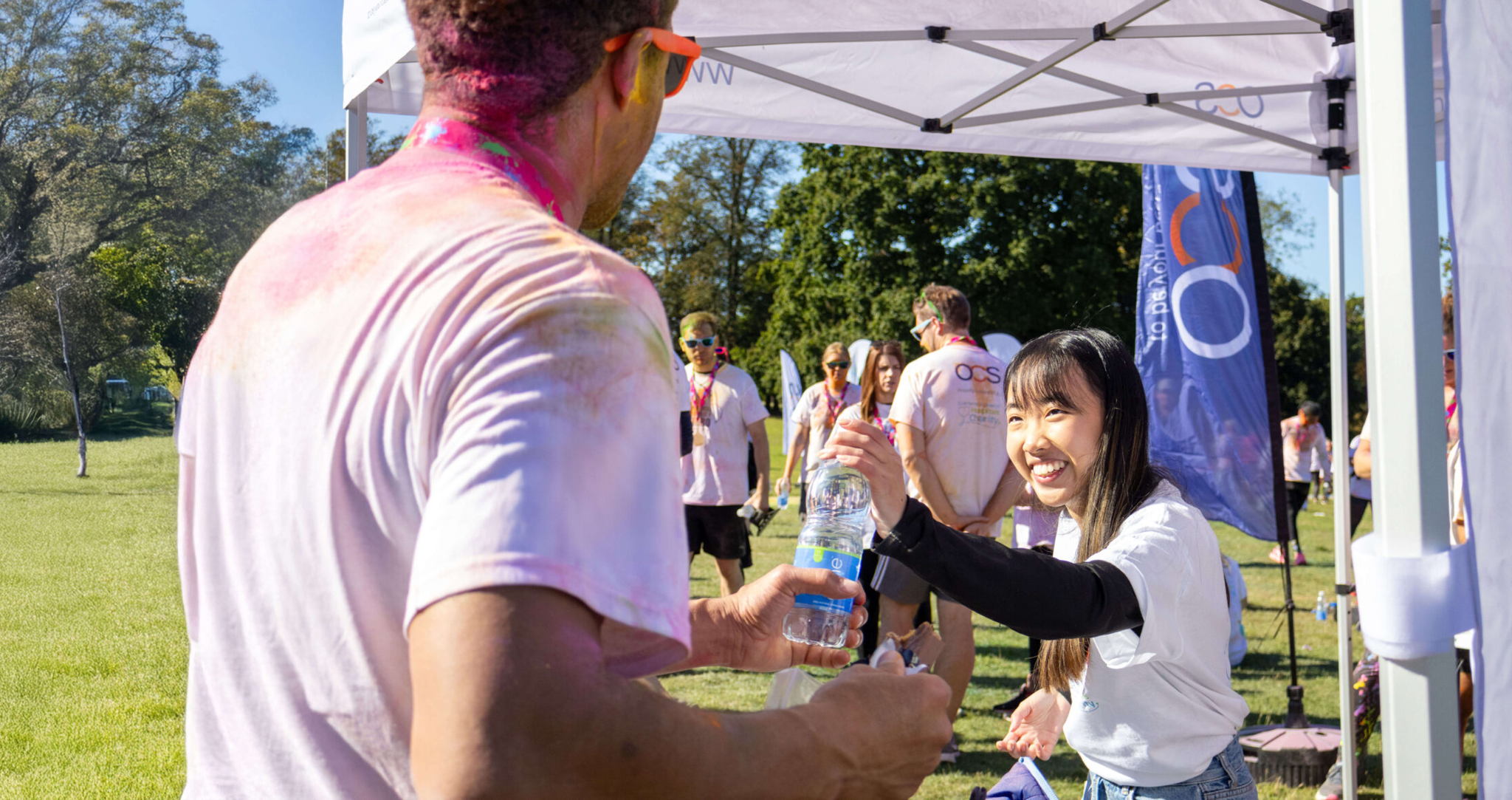 A woman smiles and hands a water bottle to a man covered in colored powder at an outdoor event, with other people and trees visible in the background on a sunny day.