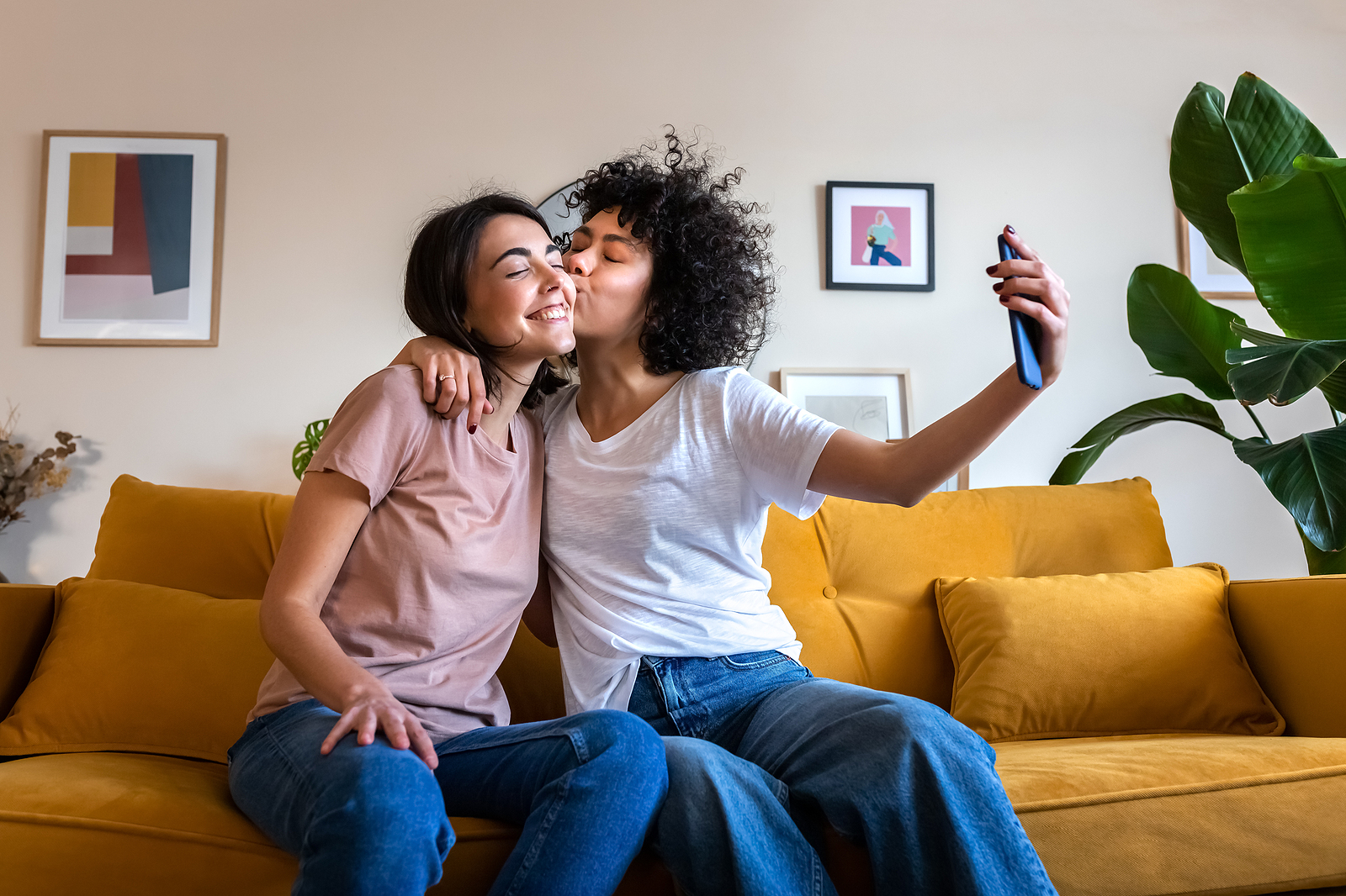 A young lesbian couple smiling sitting on their sofa taking a selfie together while sharing a kiss.