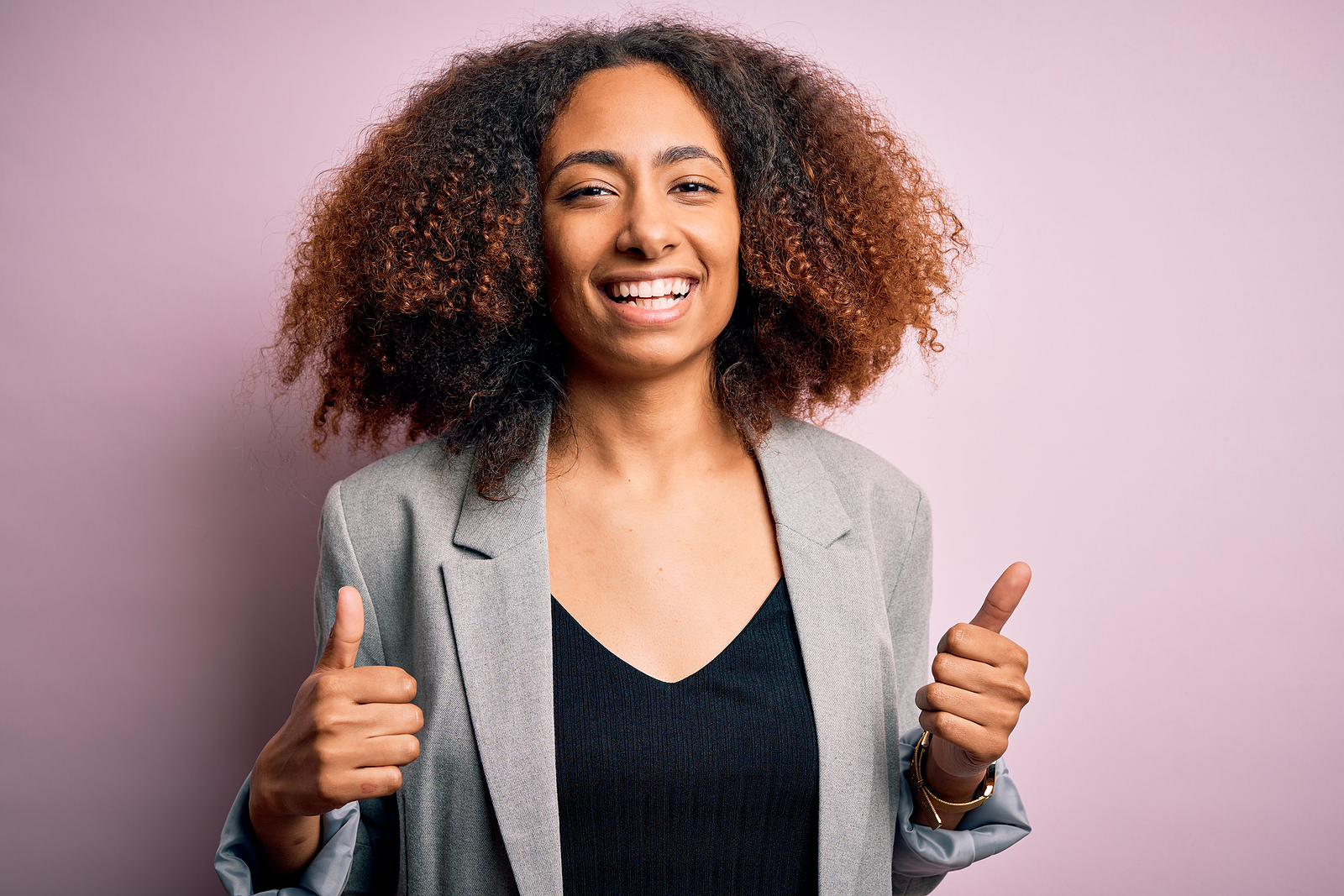 A Image of a multi ethnic attractive young woman with puffy hair and a blazer has her thumbs up in the air and smiling against a lavender background.