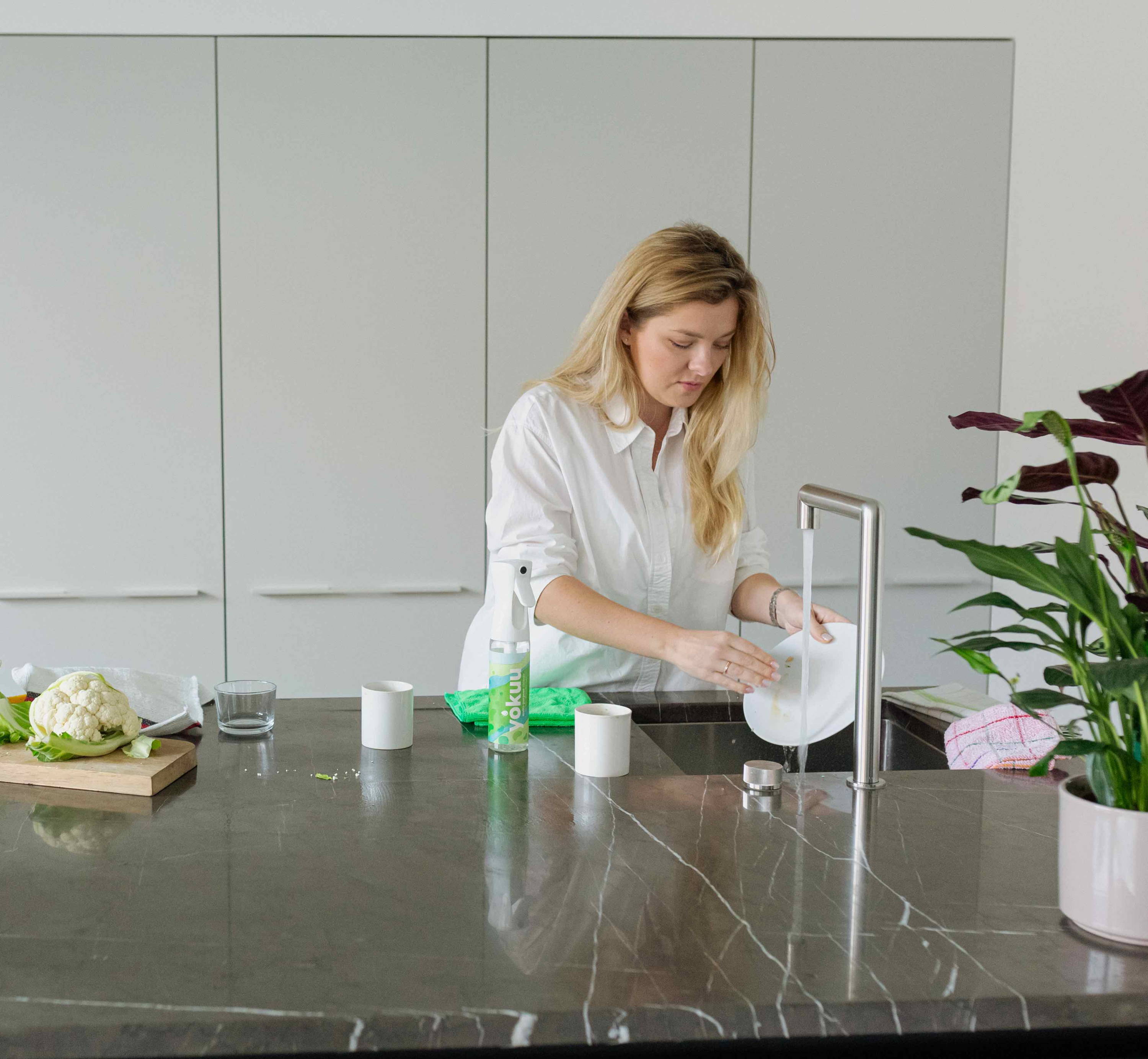 Blonde woman in a kitchen running a plate under the tap. A variety of vegetables are scattered on the counter beside her, and a bottle of Yokuu multi-purpose spray is visible in the background.