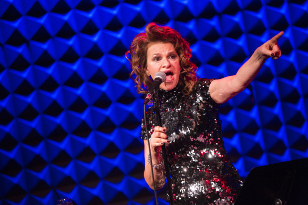 Sandra Bernhard performing a comedy act onstage. She is pointing at something out of frame while holding the mic stand.