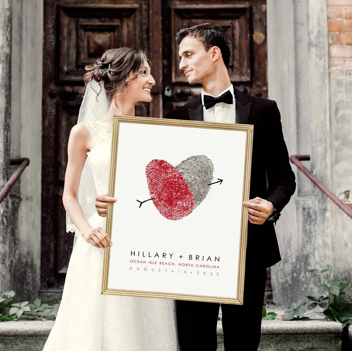Bride and groom smiling at each other, holding a framed print with overlapping red and gray fingerprint hearts. Text below reads "Hillary + Brian" and wedding details.