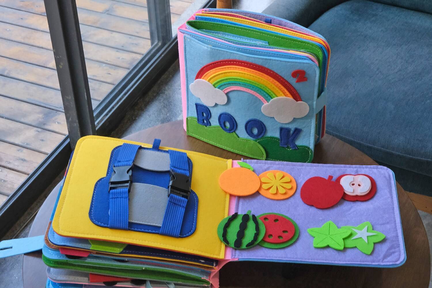 An opened educational quiet book for children showing two pages, a backpack with buckles on the left, and colorful fruits on the right.