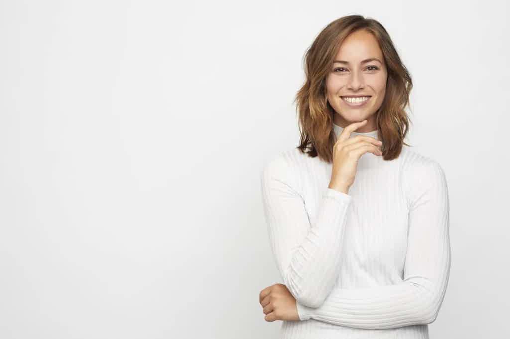 Smiling woman in a white turtleneck sweater stands against a light background, hand resting on chin.