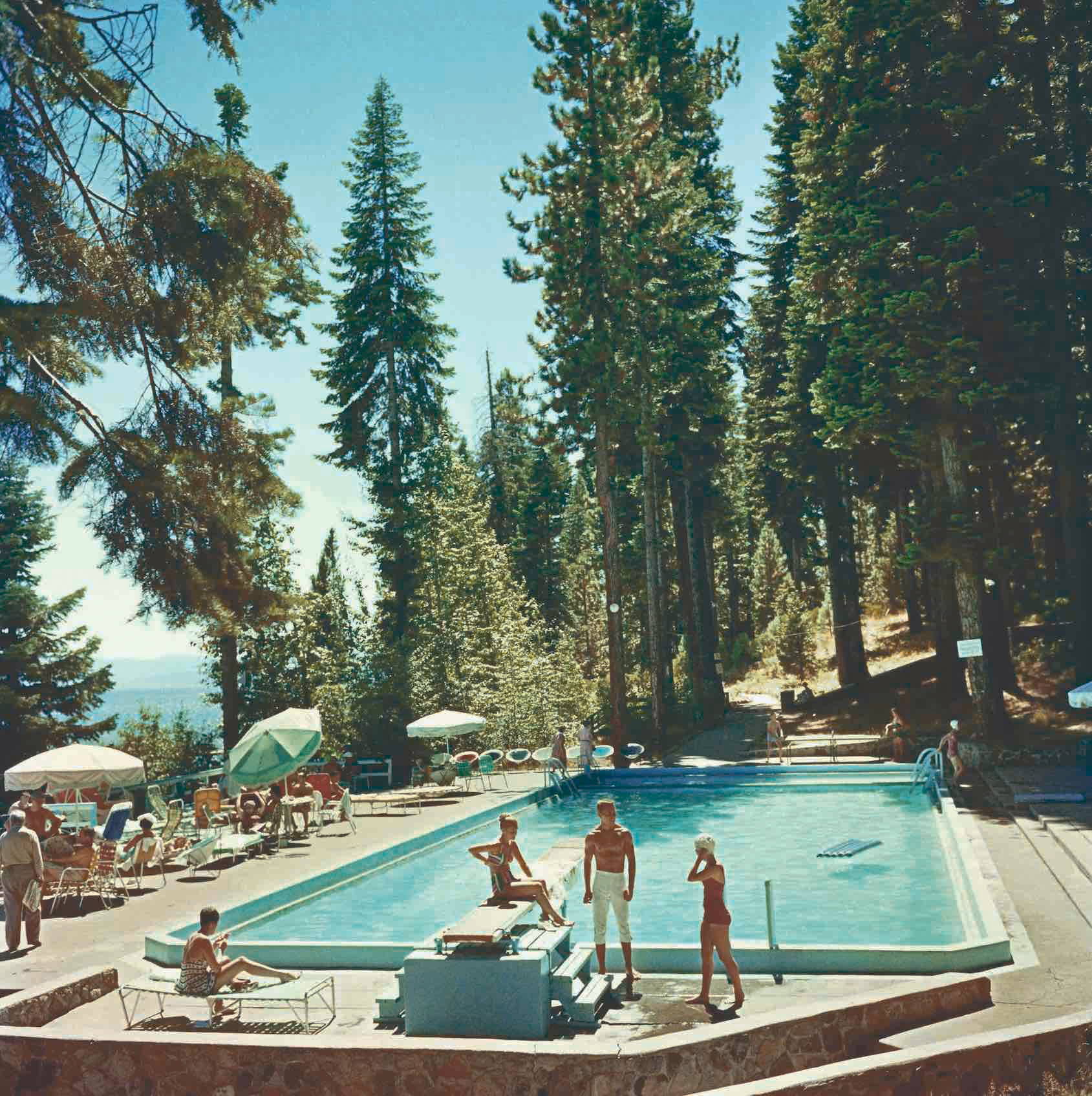 Pool At Lake Tahoe by Slim Aarons - A vintage photograph of bathers by the pool with a breathtaking backdrop of tall green trees and ocean in the distance.