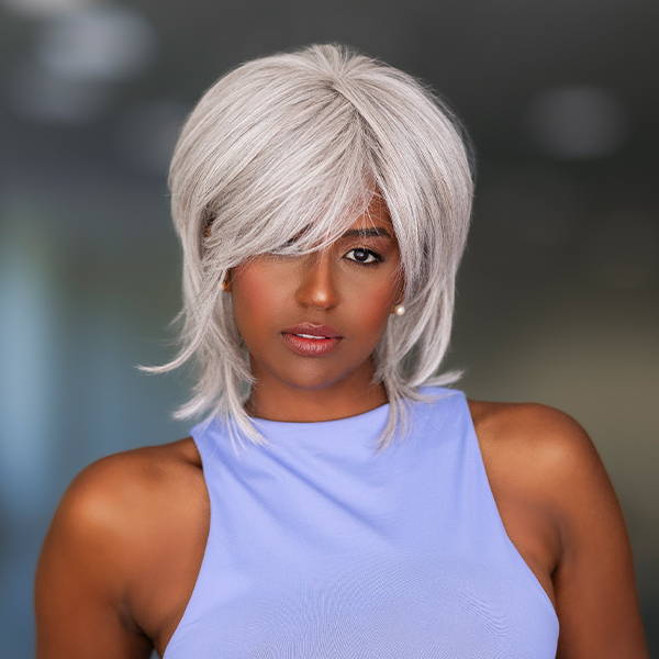 A woman wearing a wig in the style Black Tie Chic by Raquel Welch in shade RL 56/60 Silver Mist and a blue tank top is standing in a hallway with her hands in her pockets.