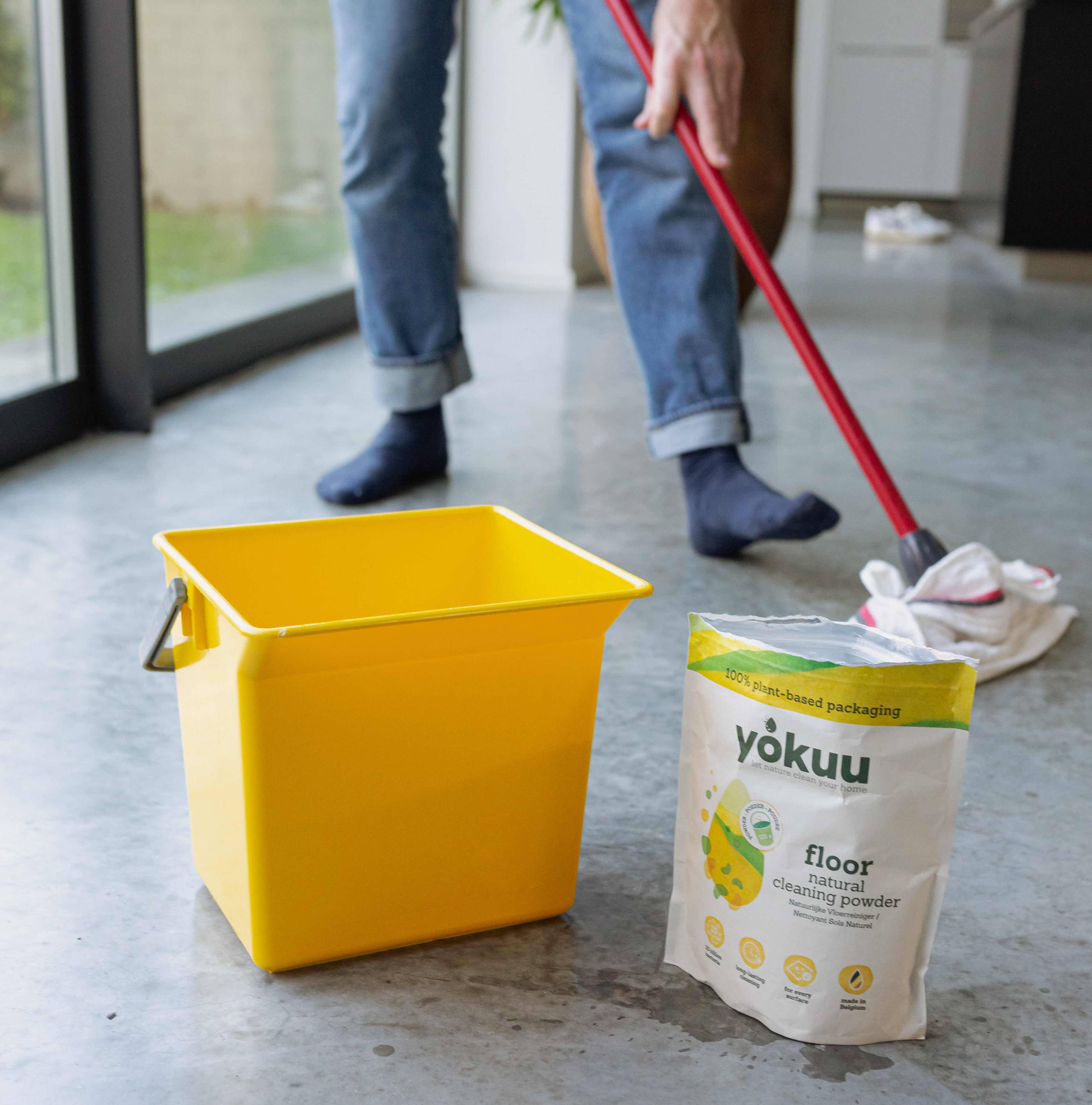 A yellow plastic bucket and an open bag of YOKUU floor cleaner are visible on a concrete floor. In the background, a pair of legs belonging to a man are visible, mopping the floor with a wet mop. The room appears to be well-lit with overhead lighting.