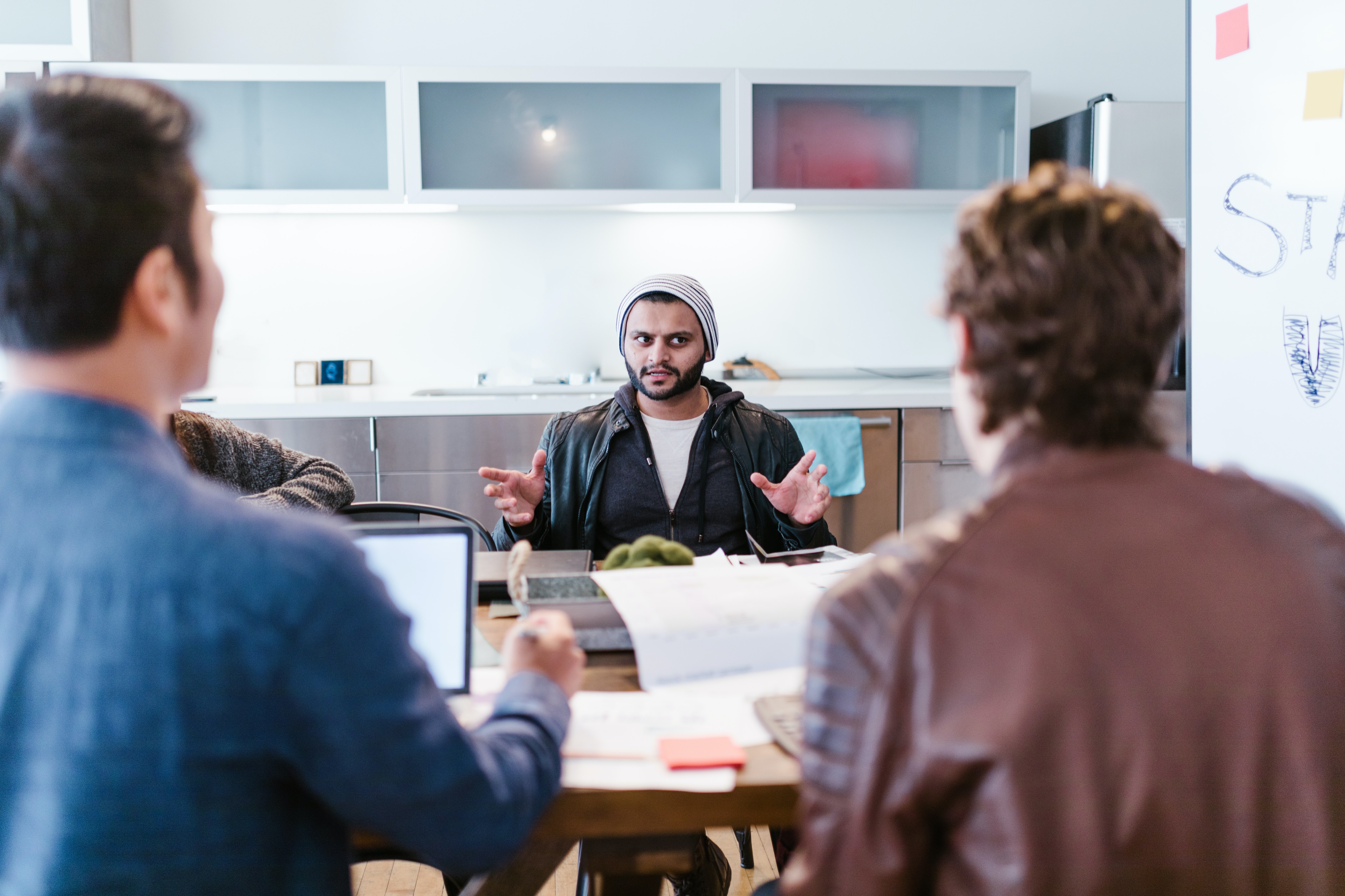 A multi ethnic man wearing a beanie and jacket talks to two friends who are doubtful of what he has to say.