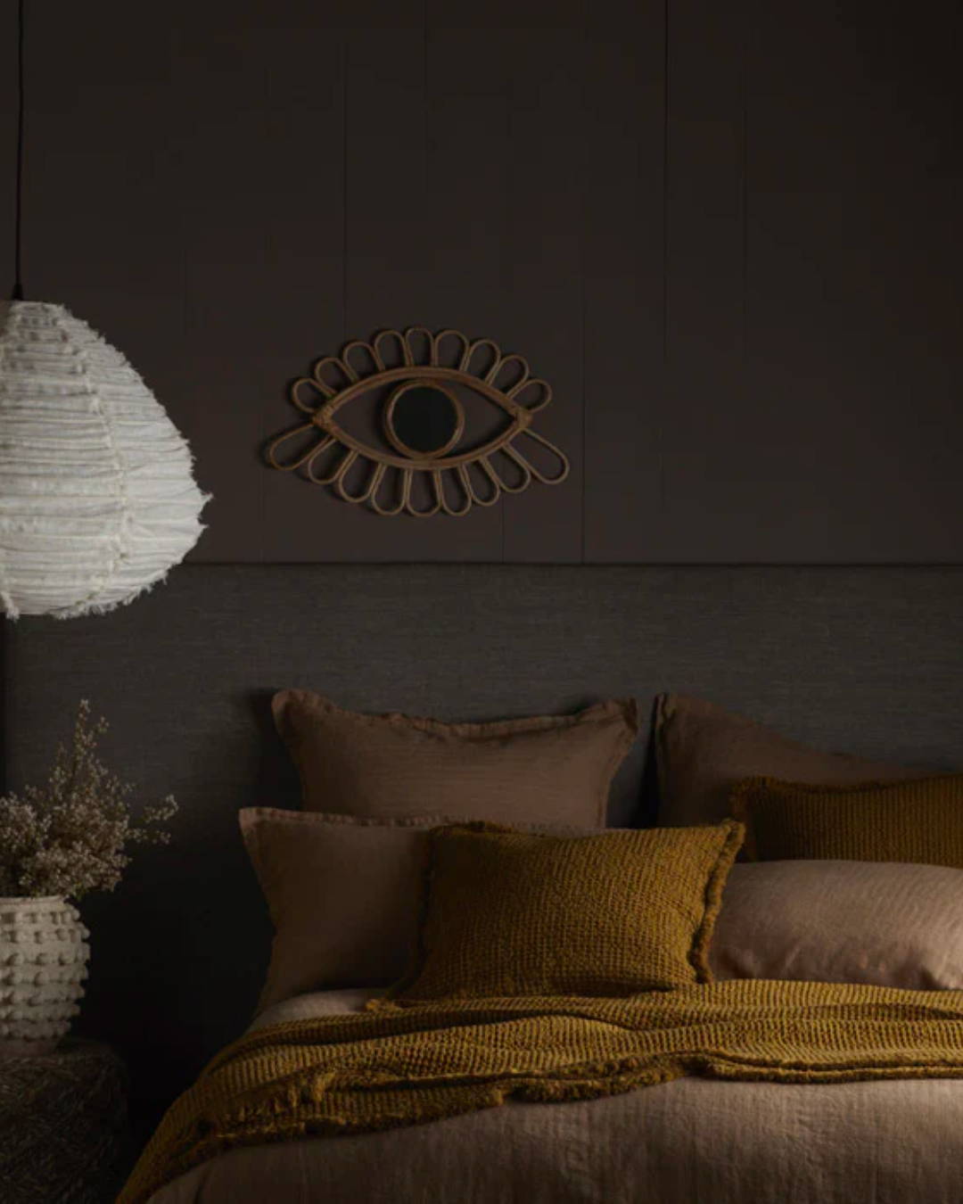 A bed made with dark linens with a waffle bedspread and matching cushion.  Behind the bed the wall is painted in a dark colour Crosby by Abigail Ahern  and a mirror in the made from bamboo in the shape of an eye. 