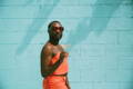 a man in an orange spaghetti strap crop top and matching pants  stands in front of a turquoise cinderblock wall for poolside pride: a slim aarons-inspired pride campaign