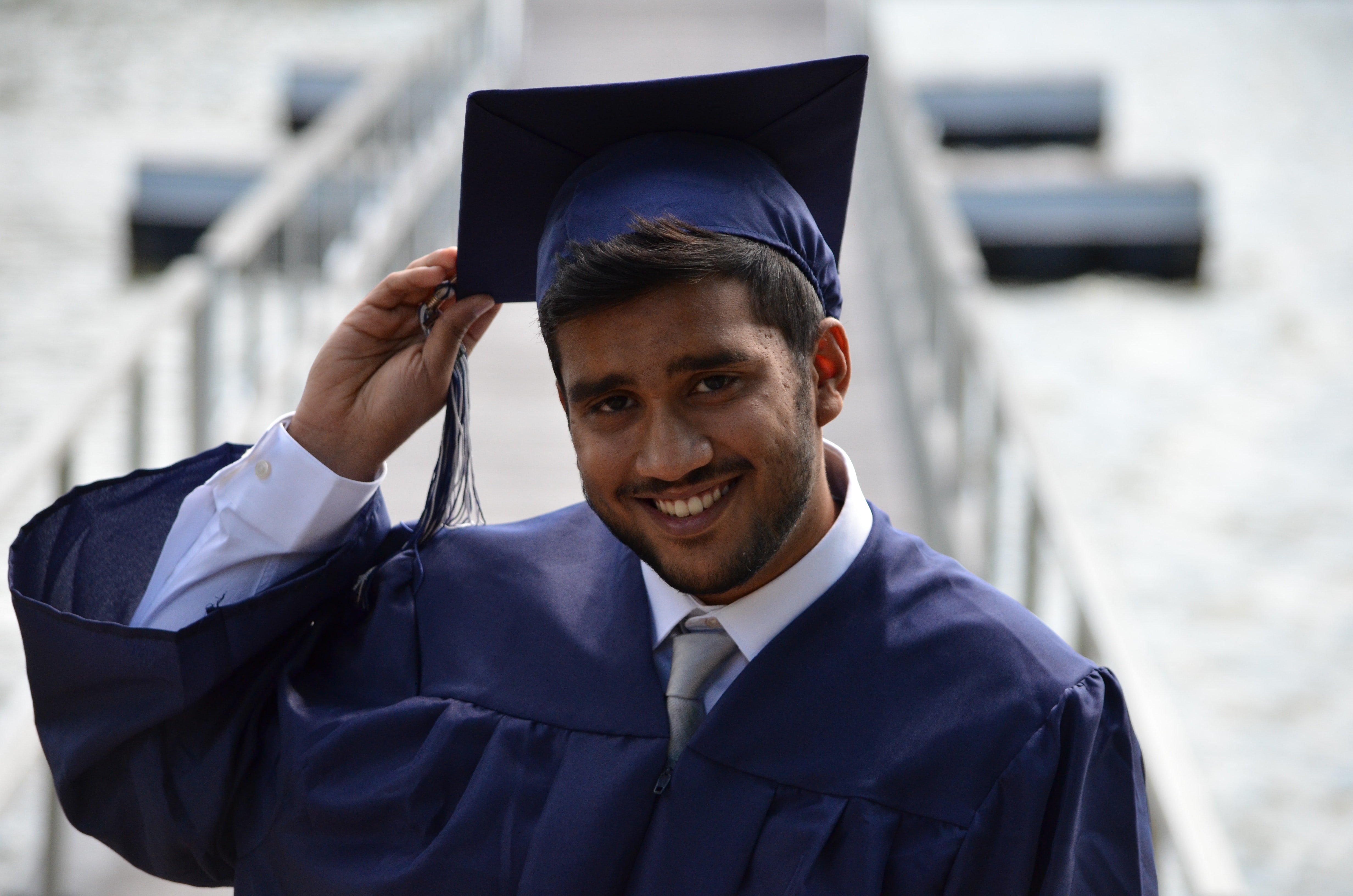 A multi ethnic man wearing a graduation cap and gown smiles and holds a hand up to his cap.