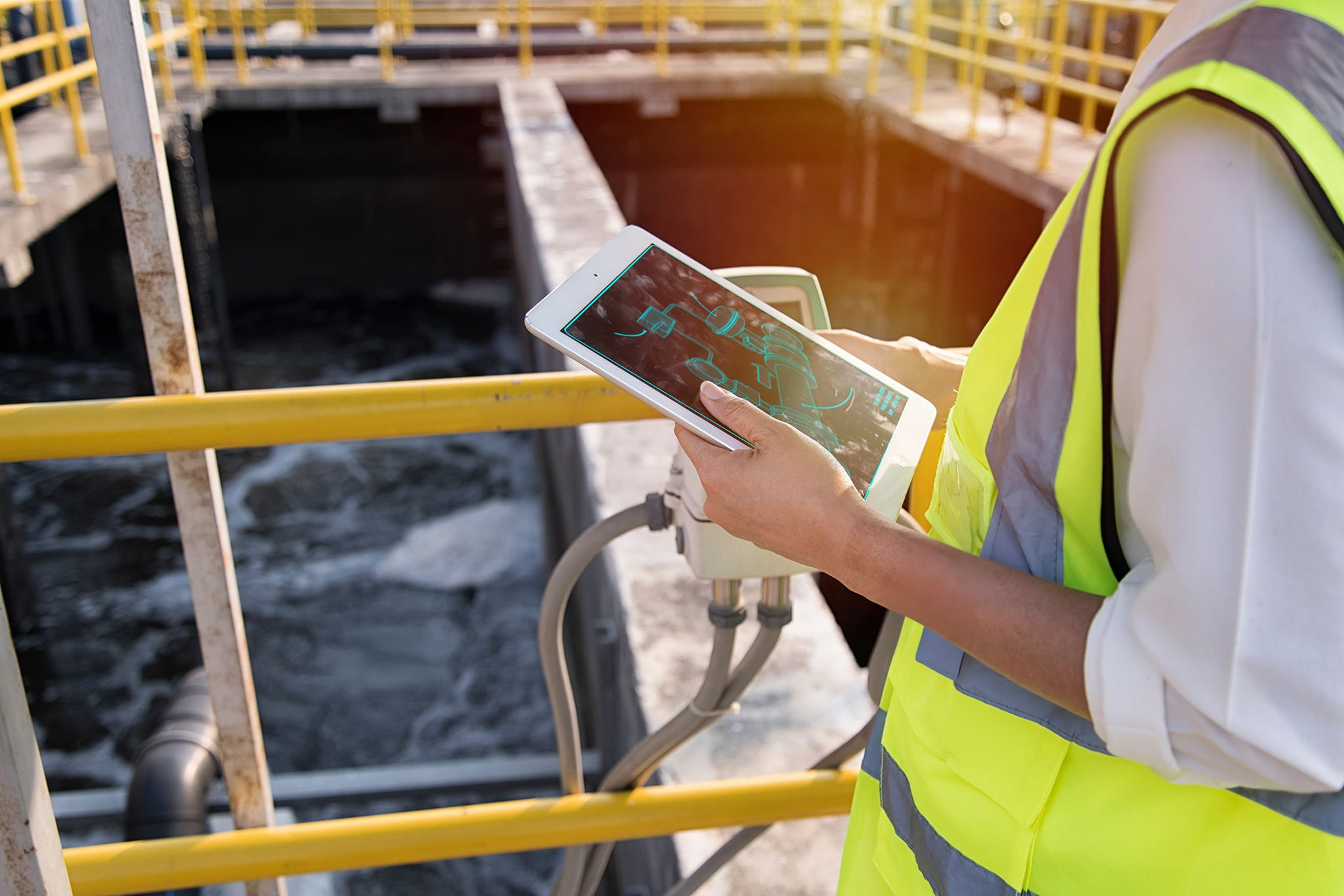 A person in a high-visibility vest uses a tablet device near railings at an industrial water treatment facility, with water and equipment visible in the background.