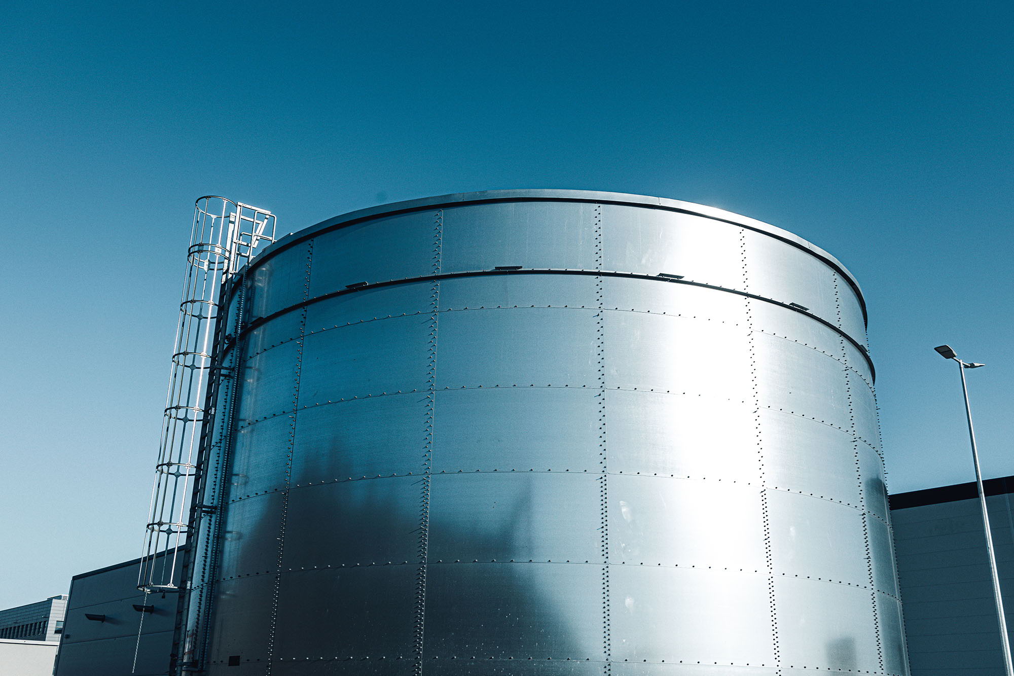 A large, shiny, cylindrical metal storage tank with a ladder on the side, set against a clear blue sky and nearby industrial buildings.