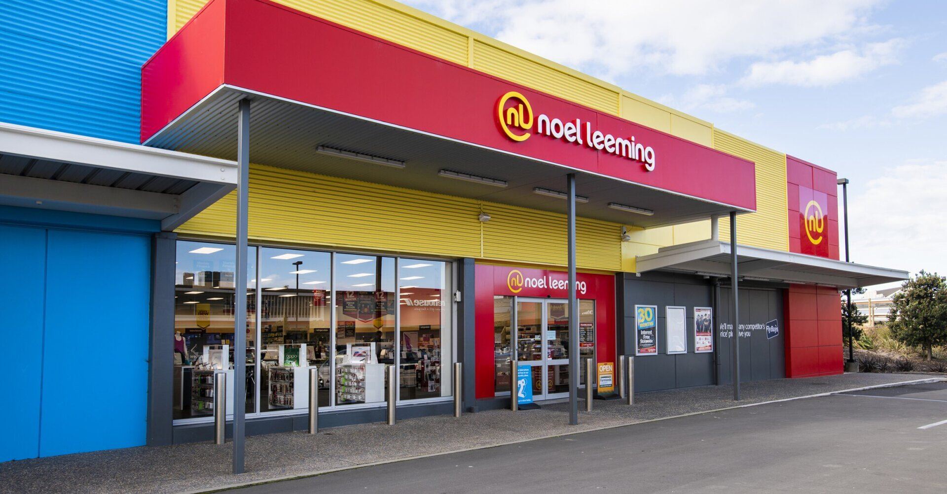 A Noel Leeming store exterior with red, yellow, and blue panels, large windows displaying electronics inside, and a clear sign above the entrance. The sky is partly cloudy.