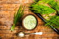 small bowl of wheat grass juice surrounded by bundles of fresh wheat grass on a wooden background