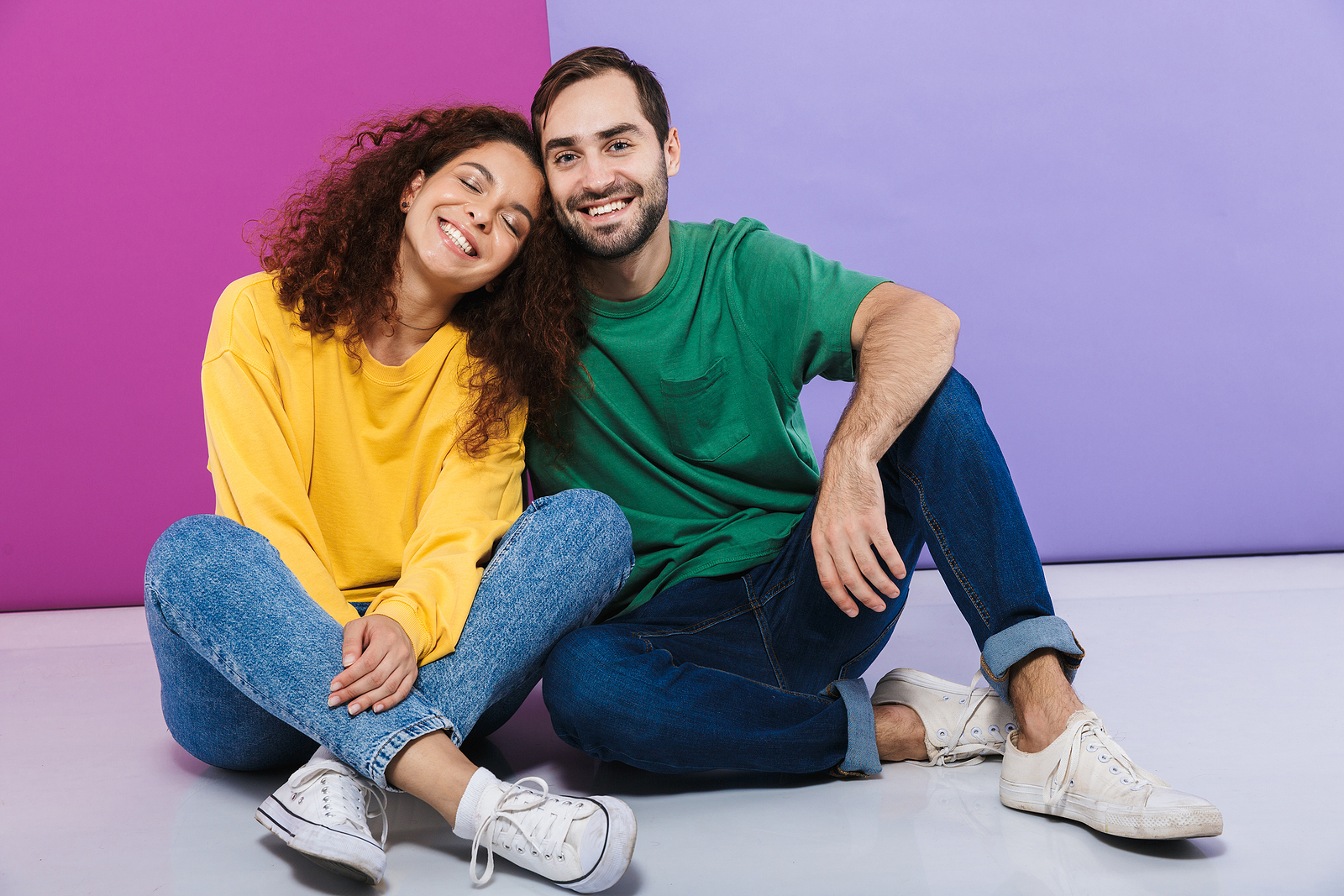 Portrait of young caucasian couple in colorful clothing smiling and hugging together while sitting on floor isolated over violet background
