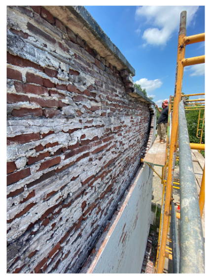 Masonry worker on scaffolding repairing a severely weathered brick wall with visible deterioration, part of a facade stabilization project.