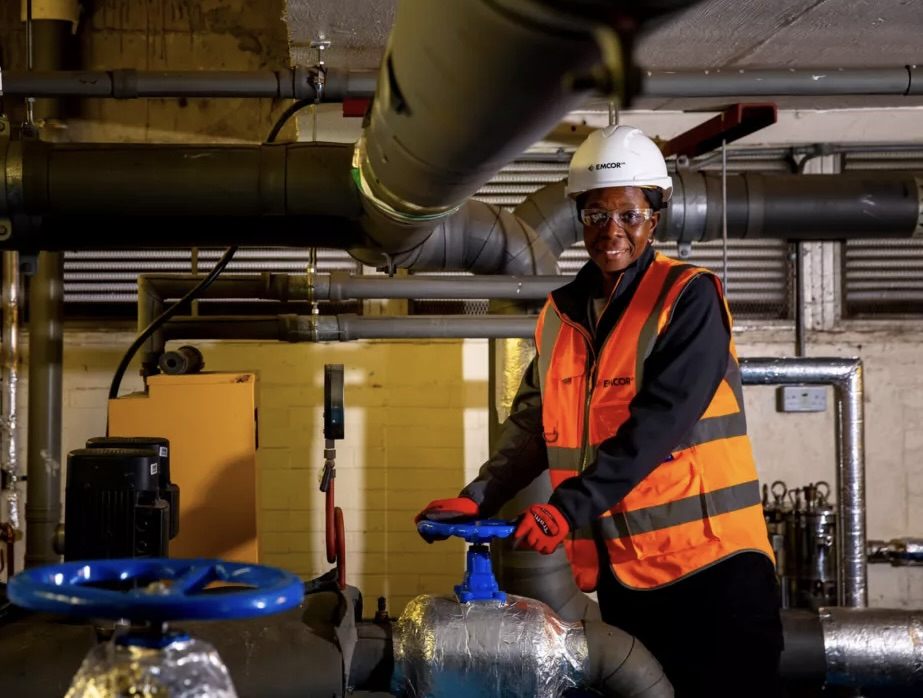 A smiling engineer wearing a white hard hat, safety glasses, orange reflective vest, and gloves operates a blue valve handle in an industrial plant with large pipes and machinery.