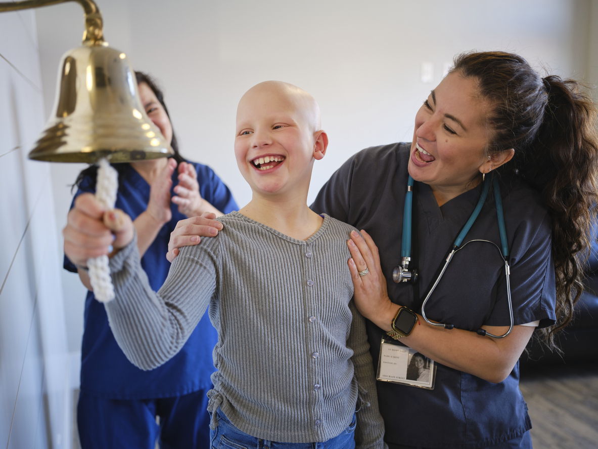 A smiling child rings a bell while a nurse with a stethoscope and another person in scrubs celebrate and clap, suggesting a joyful medical milestone.