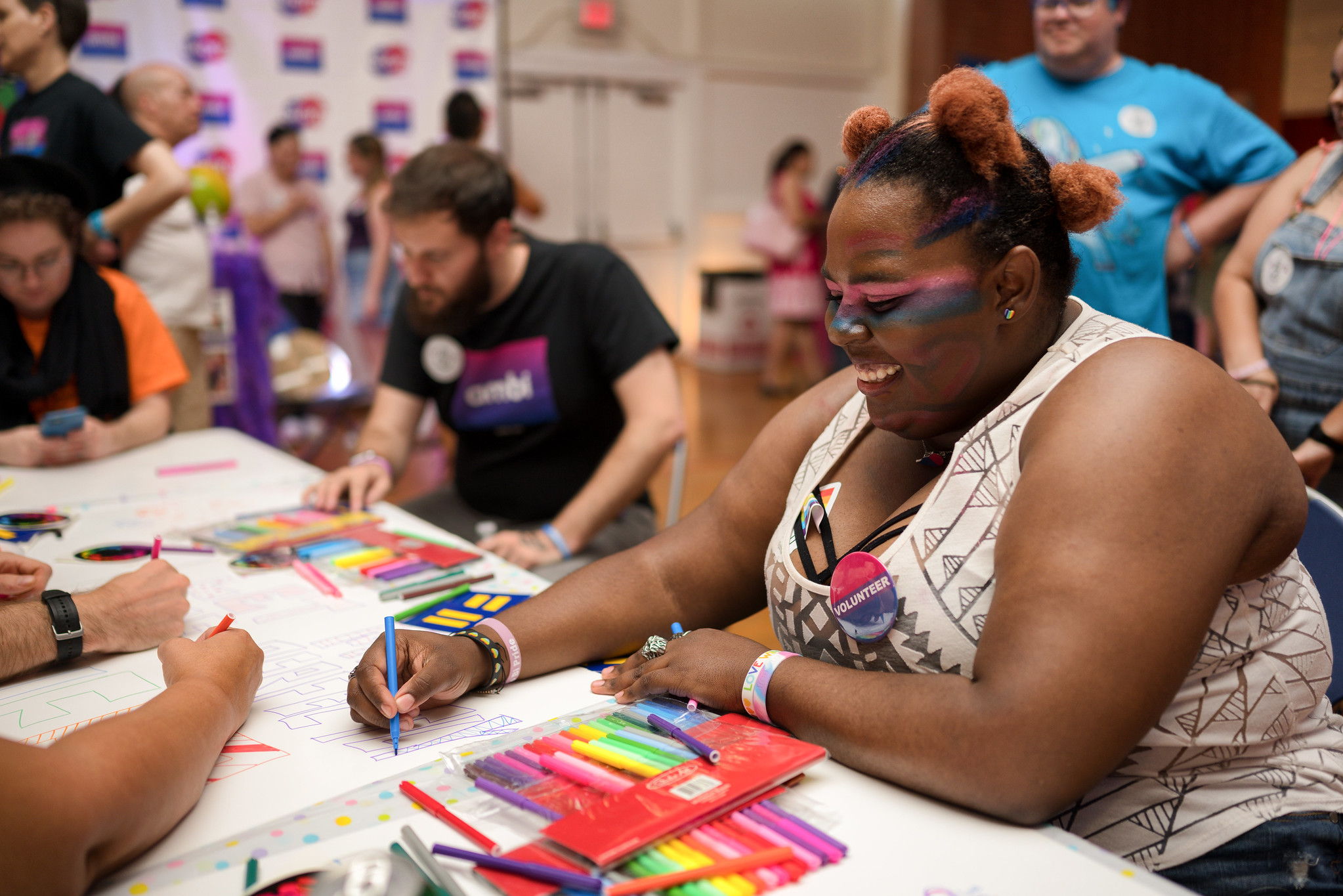 A black woman wearing a bi pin sitting at a table smiling and working on an arts craft.