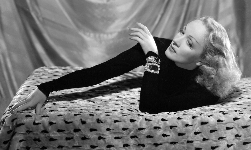 Marlene wearing a dark long sleeve shirt, laying on a bed with her hand stretched out and his hair wavy and down.