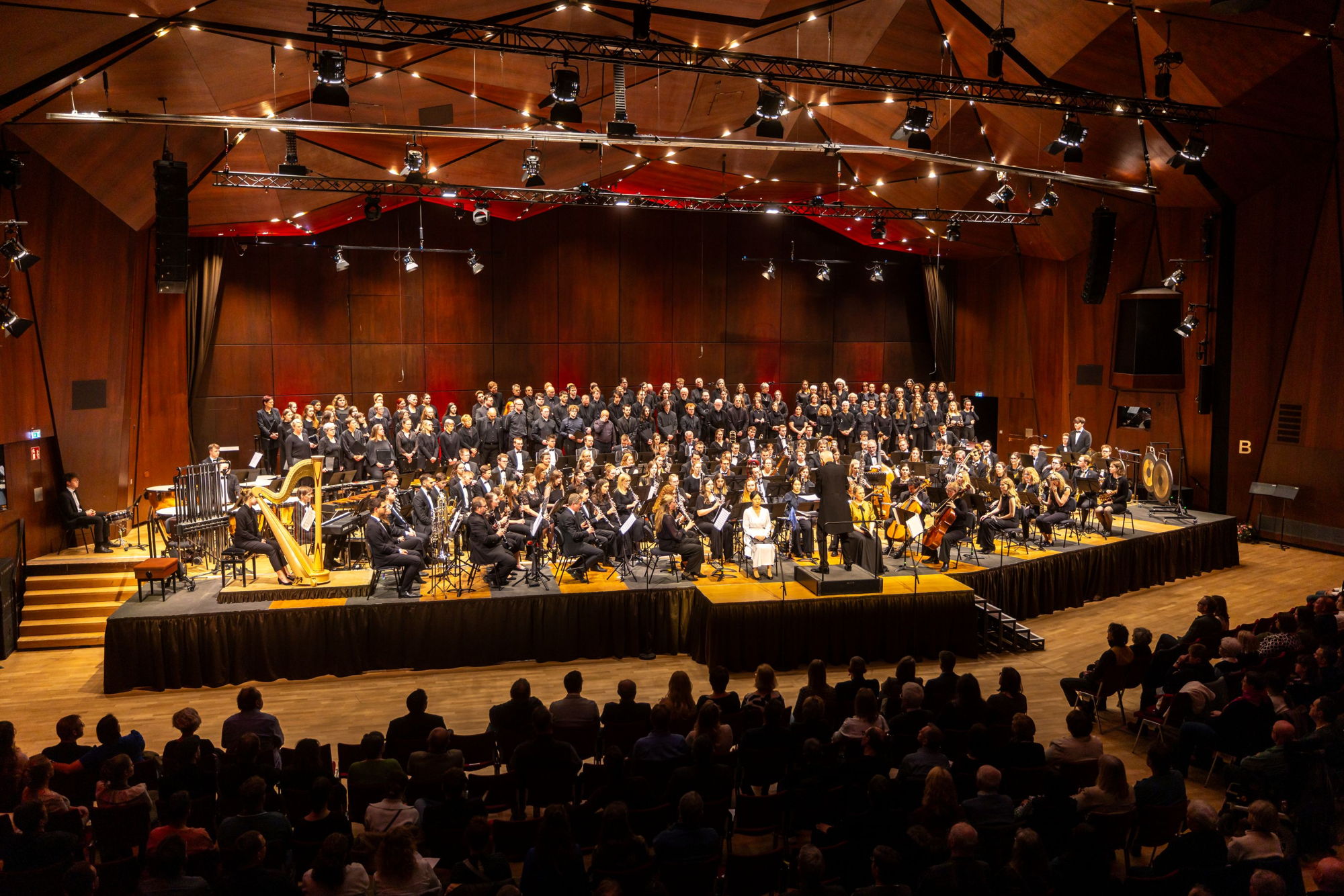Auf einer großen Bühne stehen und sitzen sehr viele Sängerinnen und Sänger sowie Blasmusikerinnen und -musiker und musizieren gemeinsam. Vor ihnen ist sehr viel Publikum zu sehen. 