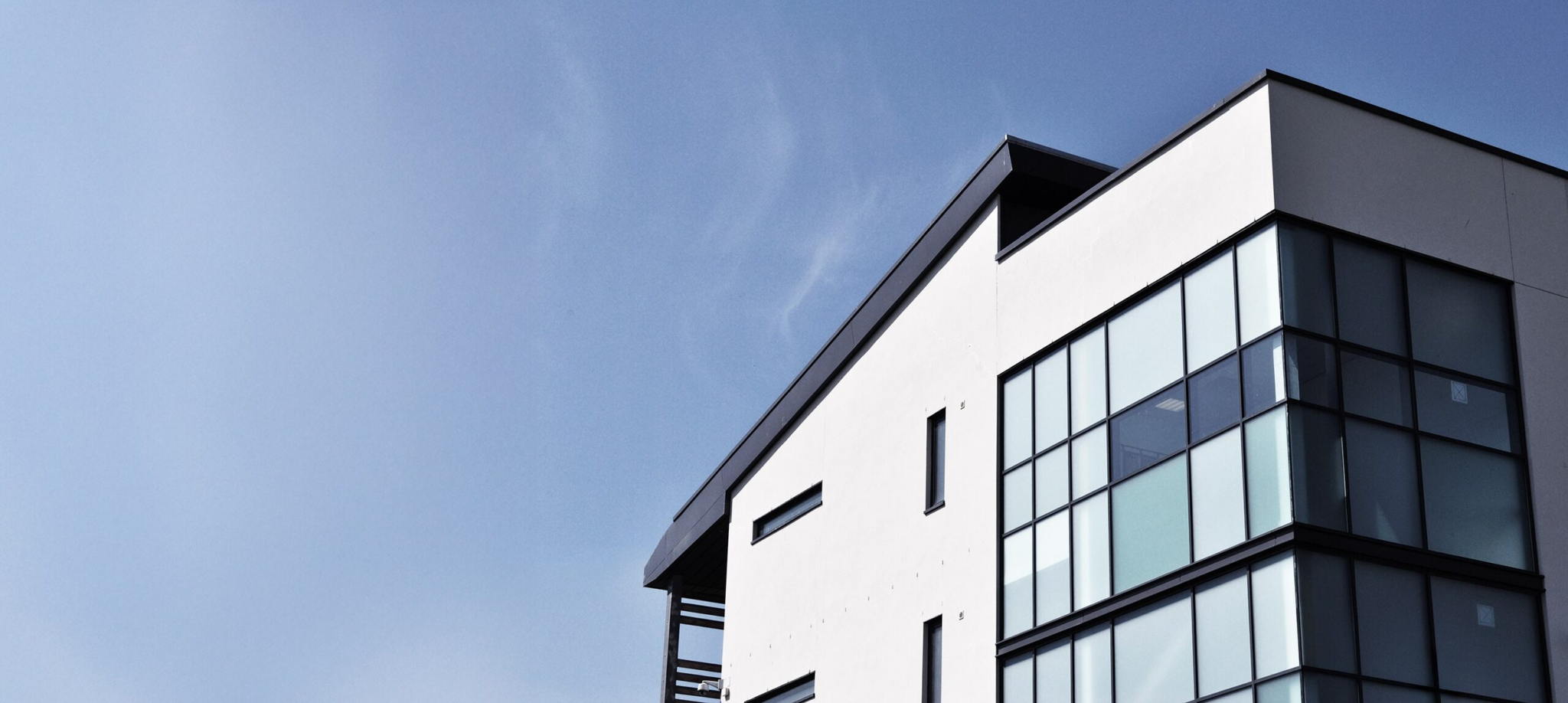 Modern white building with large glass windows and black frames, set against a clear blue sky. The architecture features clean lines and a minimalist design.