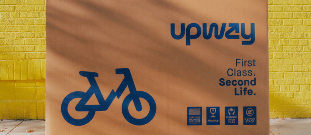 A tan Upway bike box against a yellow brick wall