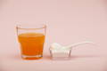 glass of orange beverage next to a spoon of white collagen powder with a pink background