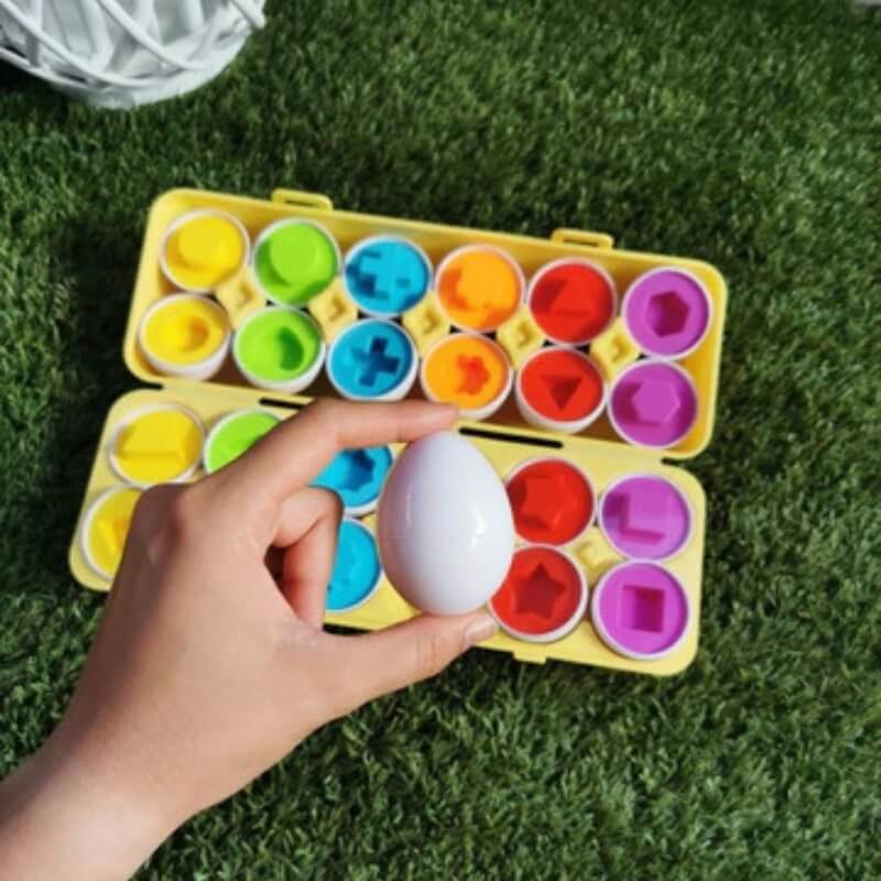 La main d'une personne peignant un œuf géométrique Montessori d'InvenToy avec un pinceau, entouré d'une palette colorée d'aquarelles posées sur de l'herbe synthétique, illustrant un jouet éducatif pour l'apprentissage des tout-petits.