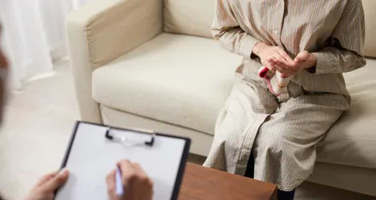 Person seated on sofa, holding a red object, while a clipboard and someone writes on a table nearby.