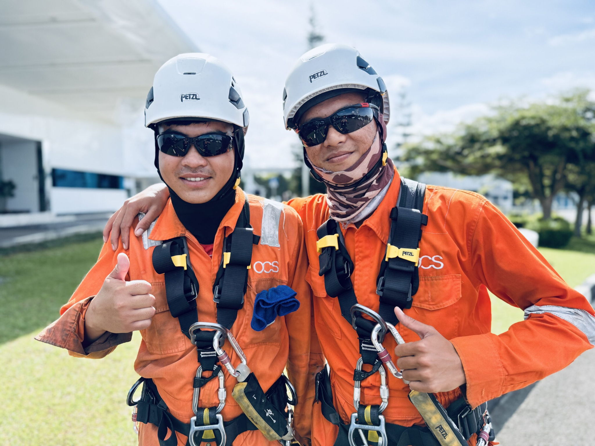 Two workers wearing orange safety uniforms, helmets, and sunglasses stand side by side outdoors, smiling and giving a thumbs-up. Safety harnesses are visible against a background of grass and a building.