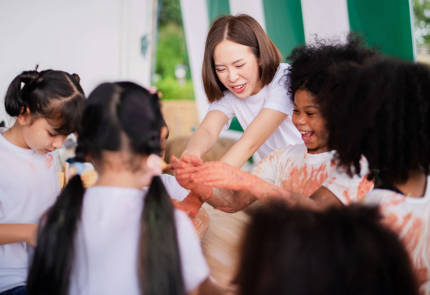 A group of young children and a woman, all wearing white shirts, smile and laugh while playing with colorful paint at a table during an arts and crafts activity.