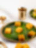 Bright orange squares of Indian sweet on a banana leaf–lined plate, with a brass lamp and cup in the background.