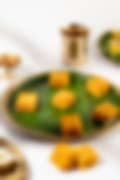 Bright orange squares of Indian sweet on a banana leaf–lined plate, with a brass lamp and cup in the background.