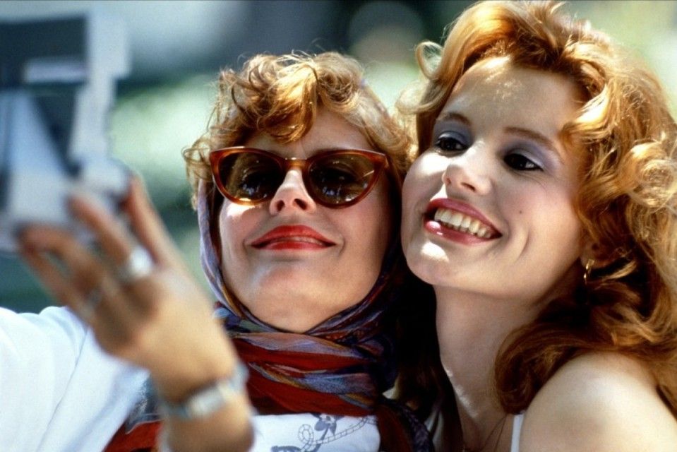 Thelma and Louise dressed up and smiling big as they take a picture of themselves on a polaroid.