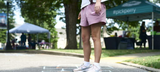 A young person plays hopscotch on pavement, evoking nostalgia, joy, and community spirit at a lively outdoor event.