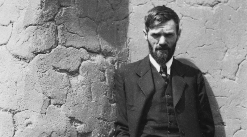 Black and white image of D.H Lawrence with a serious expression posing near a stone wall wearing a suit.
