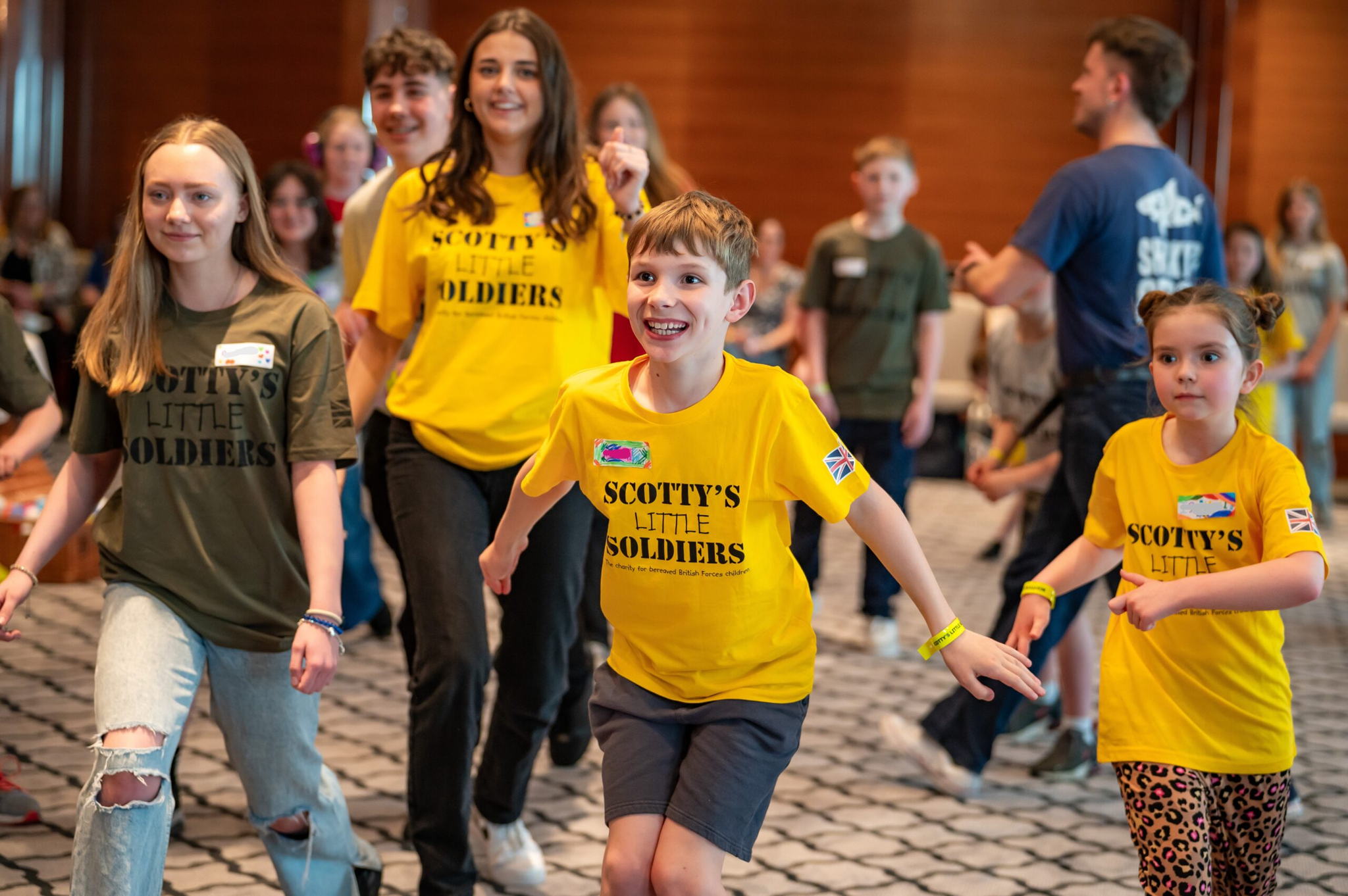A group of children and teens, some wearing yellow “Scotty’s Little Soldiers” shirts, smile and play together indoors. Adults and other children watch in the background. The mood is cheerful and lively.