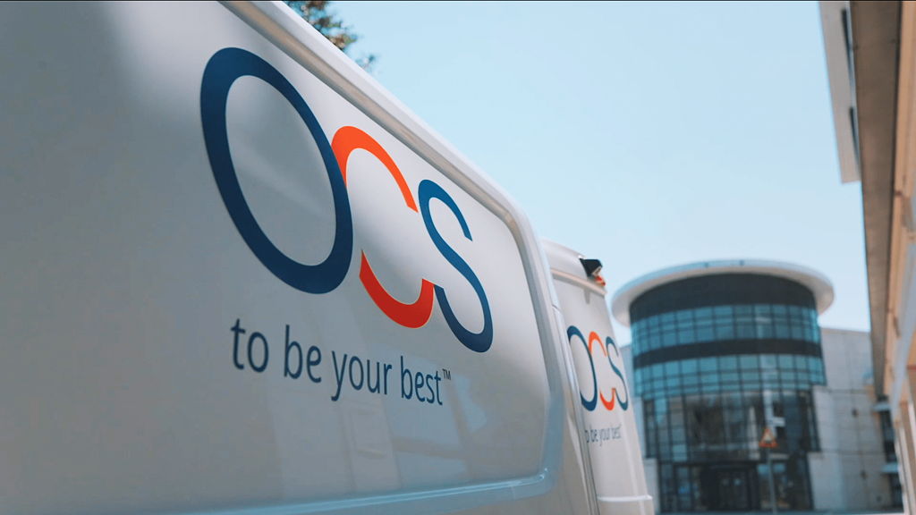 A white van with the logo OCS and the slogan to be your best is parked near a modern, circular glass building under a clear blue sky. The view is angled to capture both the van and part of the buildings facade.