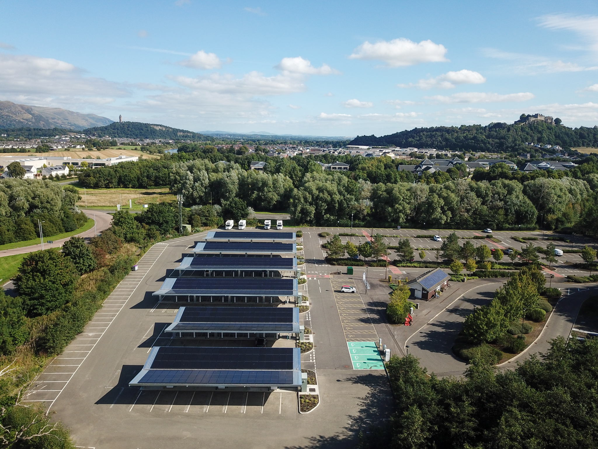 Aerial view of a large parking lot with solar panel carports, a few vehicles, and surrounding greenery. Hills and buildings are visible in the background under a partly cloudy sky.
