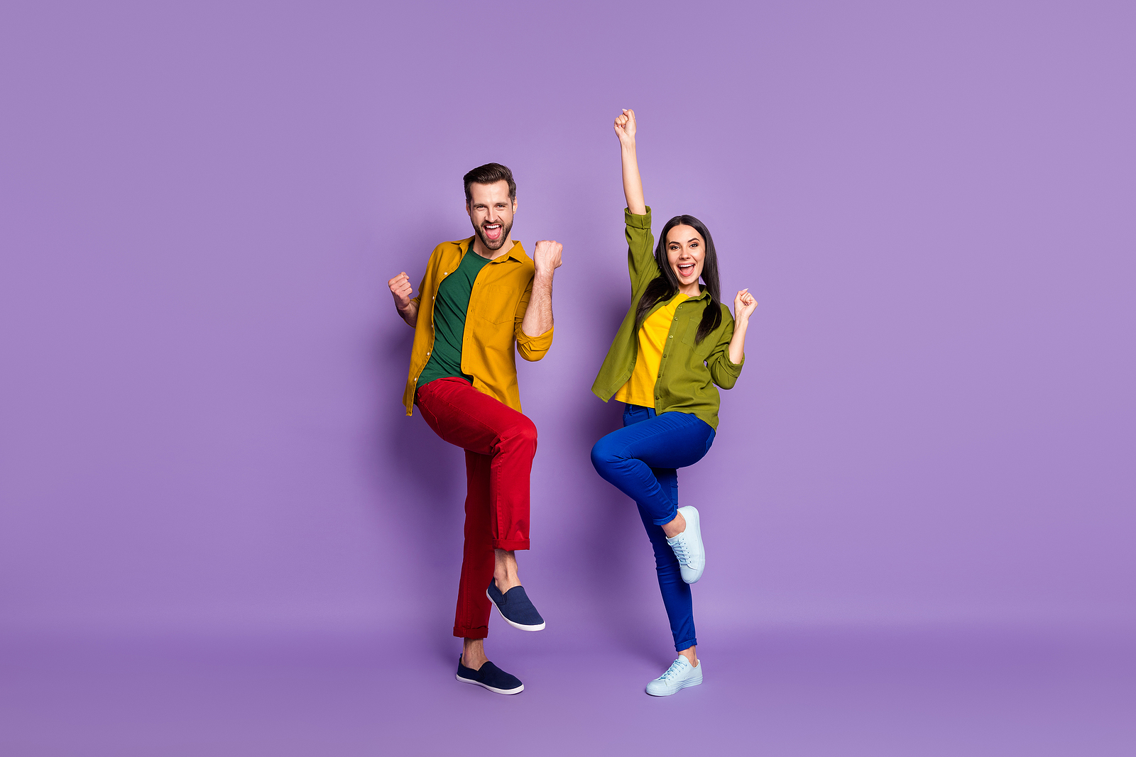 An attractive white man and woman, both wearing colorful colors against a light purple background, both are smiling and have their hands up in the air in excitement.