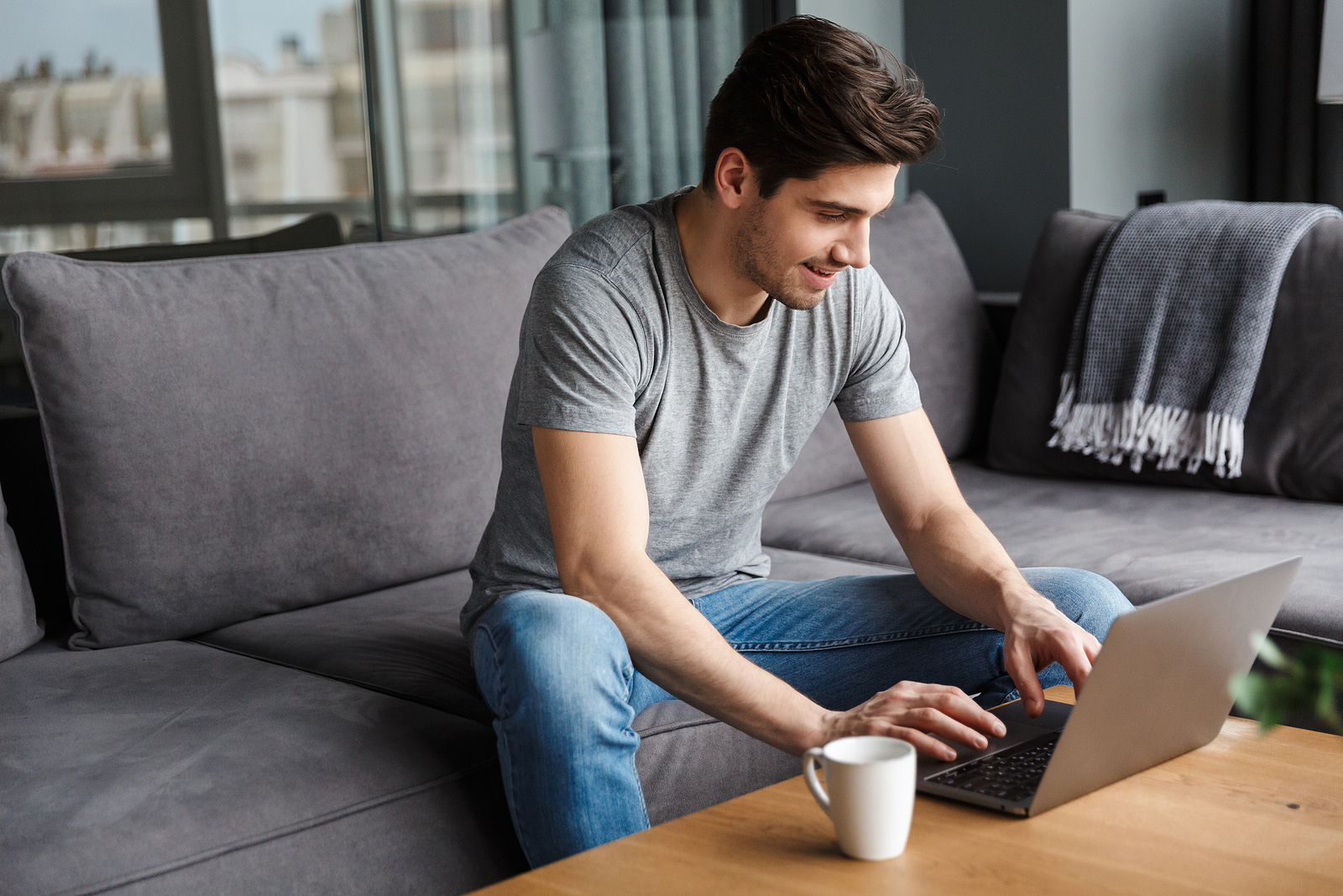 Photo of a white young man with a gray t shirt and jeans, sitting on a couch working on his laptop with a smile on his face.