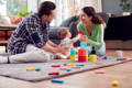 Mother, father, and their baby sitting on a carpet in the living room and playing with multicolor toys.