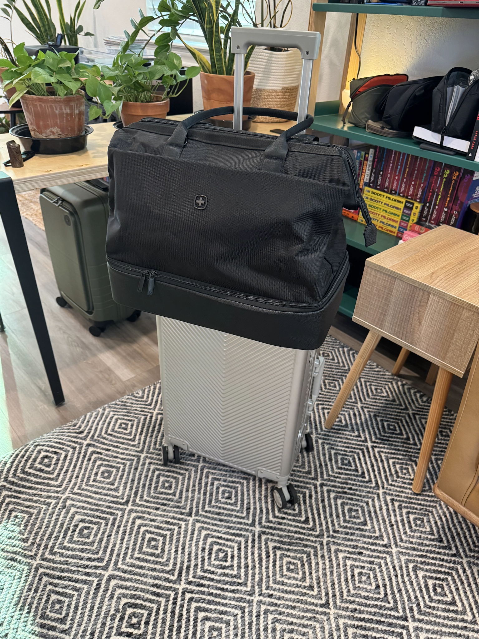 Closed black weekender bag with a zippered bottom compartment sitting on a neatly made bed in a bright, tidy room. The bag appears ready for travel, with no visible text or logos. The scene feels organized and calm, suggesting anticipation for an upcoming trip.