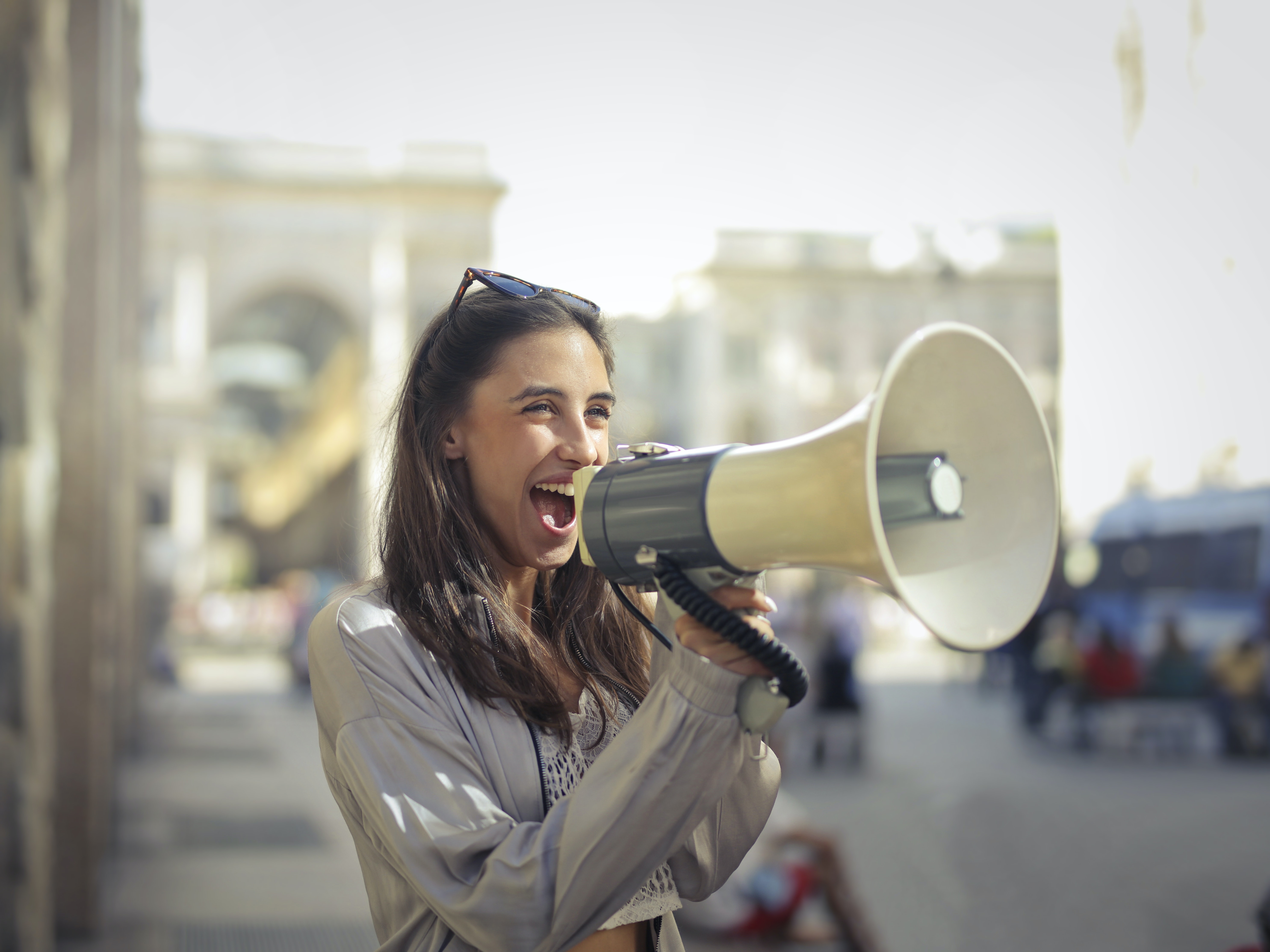 Image of a young latin woman in the street with a megaphone in her hand and she is yelling into it with a big smile on her face.