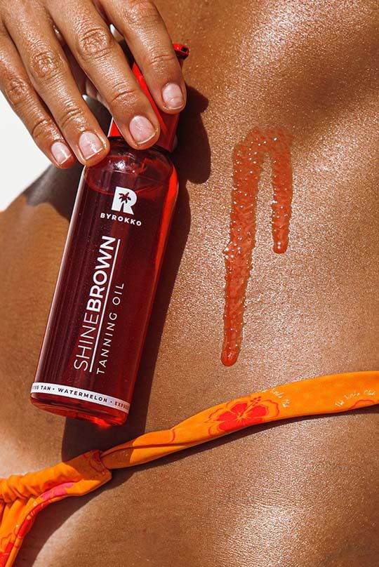 Olje Shine Brown Watermelon Oil v rokah zagorelega dekleta, in nekaj olja na njeni koži, kar prikazuje neverjetno intenzivno zagorelost, ki je posledica uporabe izdelka.