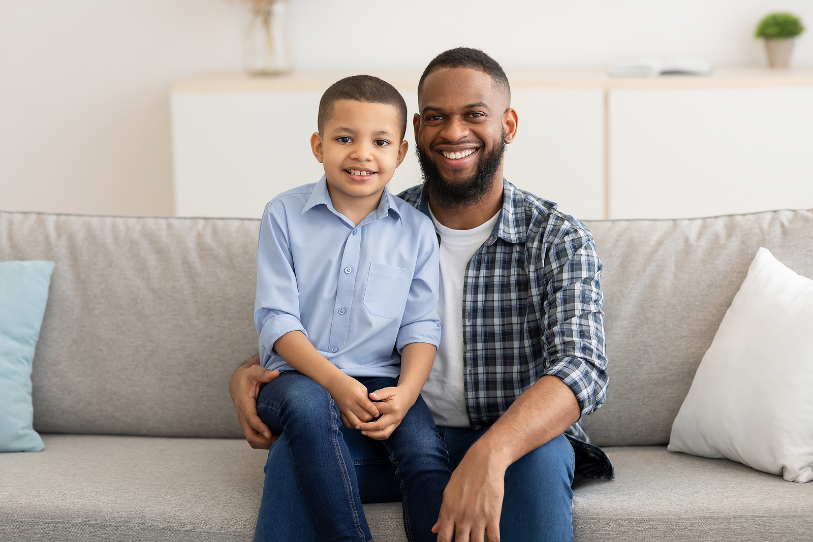 A black man sits on a couch with his young son on his lap. Both are smiling looking at the camera.