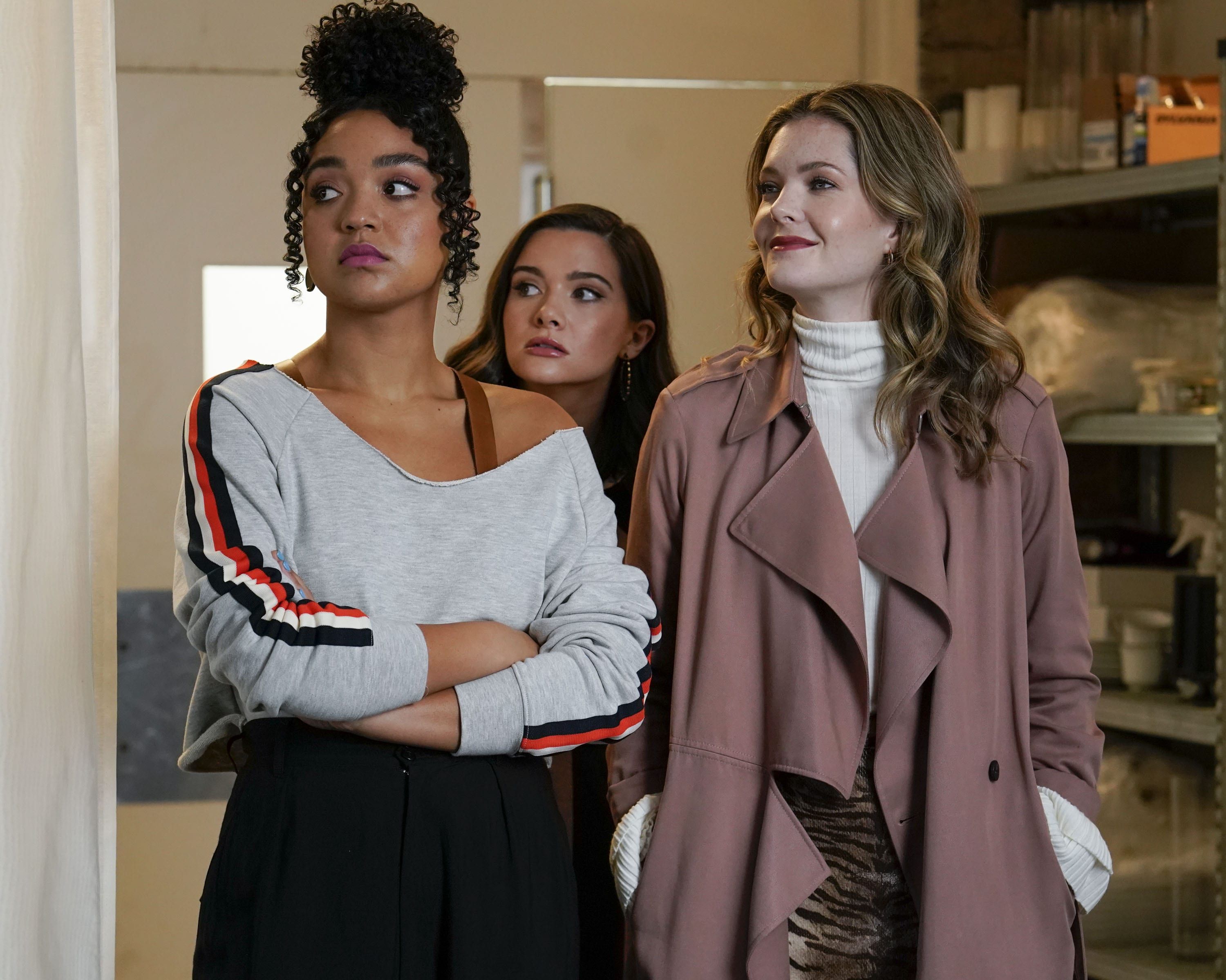 Kat, Sutton, and Jane wearing stylish warm clothing looking to their right with mixed reactions on their faces, they are in a building with supplies behind them.